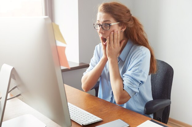 Joven estudiante pelirroja con gafas y camisa azul mirando estresado