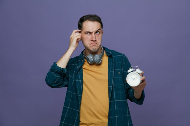 Un joven estudiante pensativo usando audífonos alrededor del cuello sosteniendo un despertador mirando a un lado mostrando un gesto de pensar aislado en un fondo morado