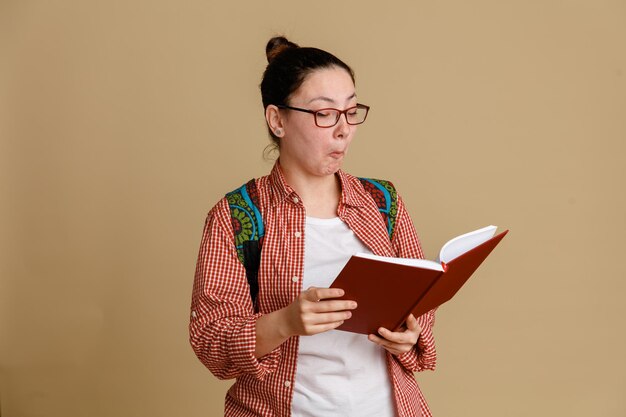 Joven estudiante con ropa informal y gafas con mochila sosteniendo un cuaderno que parece intrigado leyendo algo de pie sobre un fondo marrón