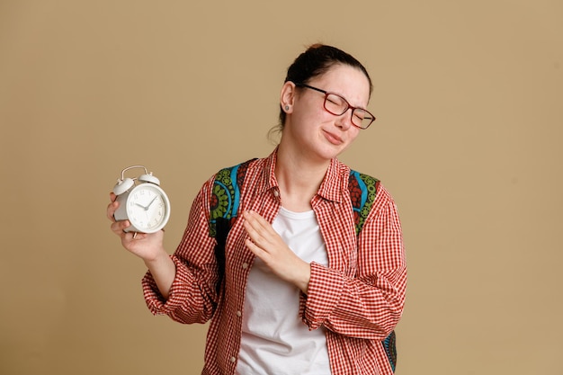 Joven estudiante con ropa informal y gafas con mochila sosteniendo un despertador que parece confundida y preocupada haciendo un gesto de alto con la mano parada sobre fondo marrón