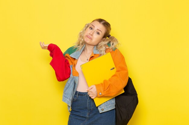 joven estudiante en ropa moderna sosteniendo archivos amarillos y posando en amarillo