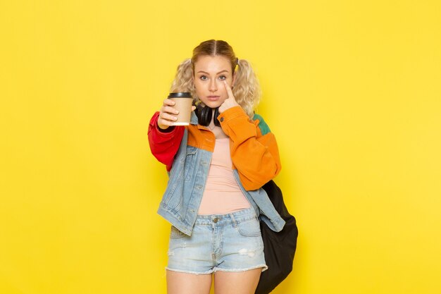 joven estudiante en ropa moderna sosteniendo café y posando en amarillo