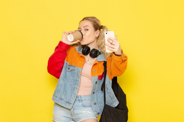 Joven estudiante en ropa moderna tomando un selfie con café en amarillo