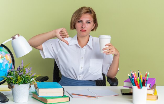 Joven estudiante rubia disgustada sentada en un escritorio con herramientas escolares sosteniendo una taza de café de plástico mostrando el pulgar hacia abajo