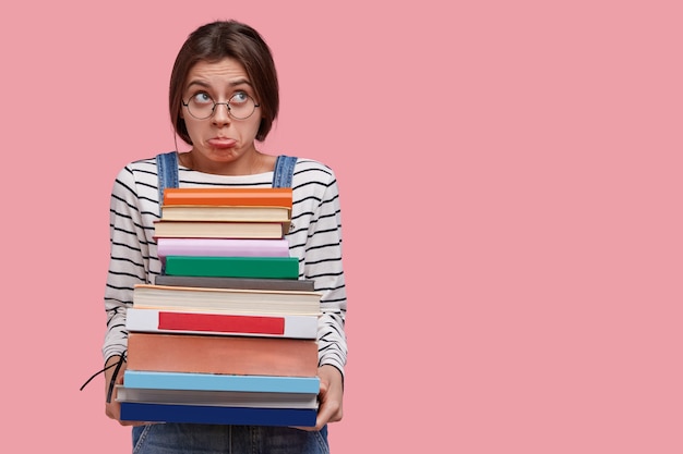 Joven estudiante universitario disgustado caucásico sostiene pila de libros, se prepara para la sesión de examen, frunce los labios en descontento