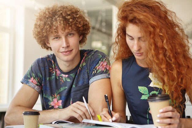 Joven estudiante universitario sentado cerca de su amiga pelirroja que lo está ayudando a estudiar