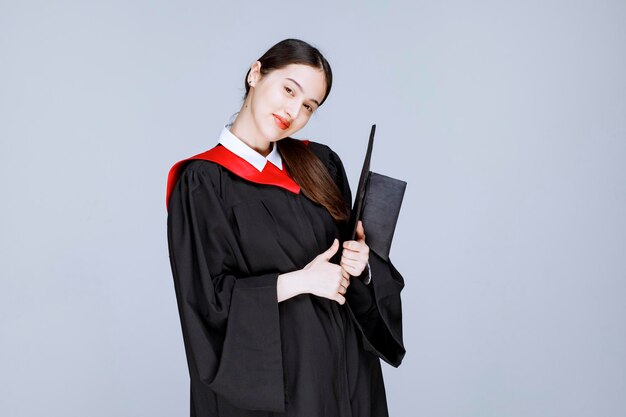 Joven estudiante en vestido sosteniendo gorro y posando. Foto de alta calidad