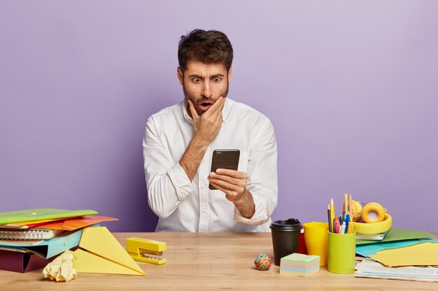 Joven estupefacto lee un mensaje de texto, mira fijamente con mirada asustada, sostiene un teléfono móvil moderno, trabaja en la oficina, viste una elegante camisa blanca