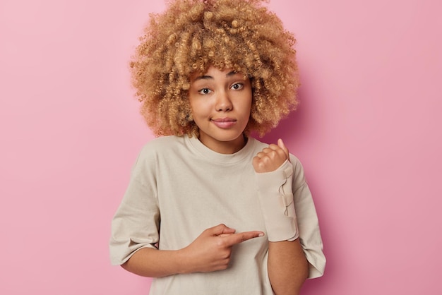 Una joven europea de cabello rizado muestra un brazo roto después de un accidente y usa una venda elástica vestida con una camiseta informal aislada sobre un estudio rosado Concepto de accidente de seguro y atención médica