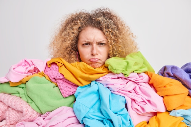 La joven europea descontenta con el pelo rizado tiene una expresión de mal humor sobrecargada con ropa colorida molesta por tener mucho trabajo doméstico tiene que doblar la ropa lavada aislada sobre una pared blanca