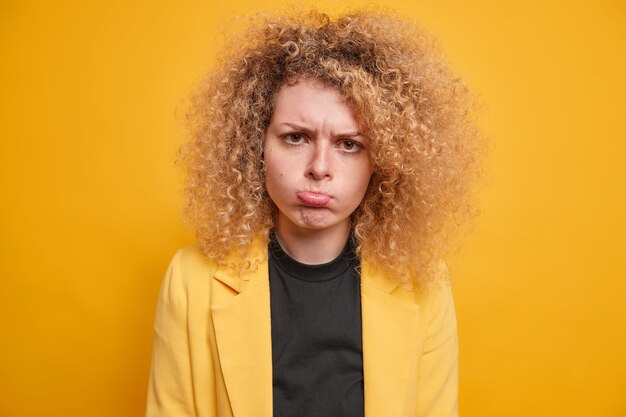 La joven europea disgustada con el pelo rizado natural se frunce los labios y se siente frustrada al estar decepcionada por algo que hace que la mueca ofendida infeliz use un traje formal amarillo que se encuentra en el interior