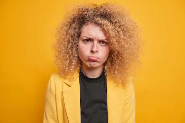 La joven europea disgustada con el pelo rizado natural se frunce los labios y se siente frustrada al estar decepcionada por algo que hace que la mueca ofendida infeliz use un traje formal amarillo que se encuentra en el interior