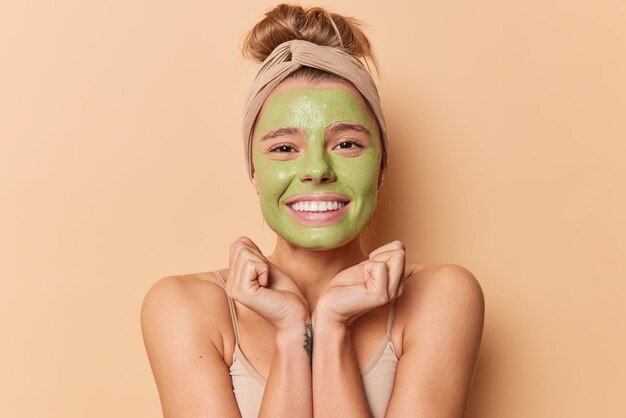 Una joven europea feliz mantiene las manos bajo las sonrisas de la barbilla con gusto aplica una máscara nutritiva verde que usa una banda para la cabeza se somete a procedimientos de belleza para el tratamiento de la piel aislado sobre un fondo beige.