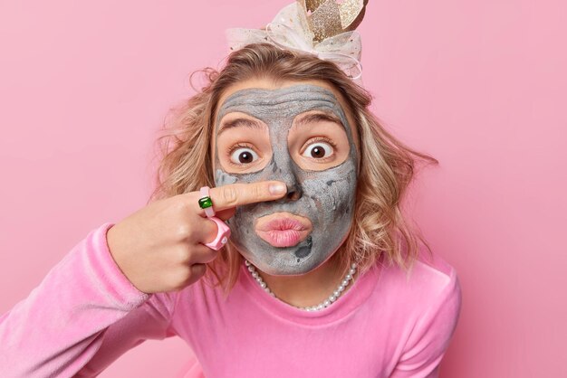 Una joven europea maravillada con peinado aplica una máscara de arcilla hidratante en la cara toca la nariz tiene los ojos muy abiertos lleva puente de corona y collar aislado sobre fondo rosa Concepto de belleza