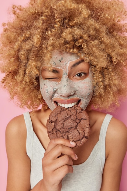 Una joven europea positiva con el pelo rizado y tupido muerde una apetitosa galleta de chocolate guiña el ojo aplica una máscara de belleza en la cara vestida de forma informal cuida la piel aislada sobre un fondo rosa