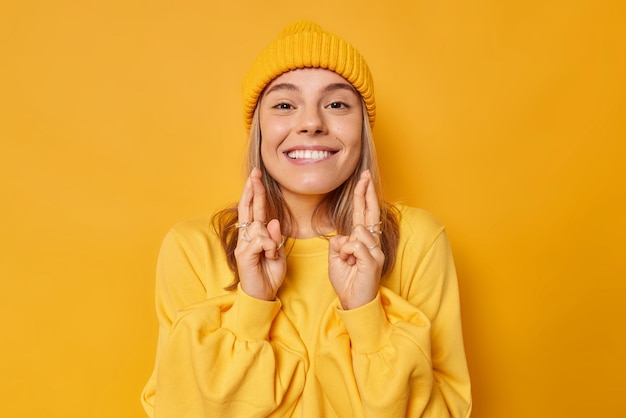 Una joven europea positiva sonríe alegremente con los dientes, mantiene los dedos cruzados, cree en la buena suerte, reza, desea fortuna, usa sombrero y puente aislado sobre fondo amarillo. Concepto de lenguaje corporal