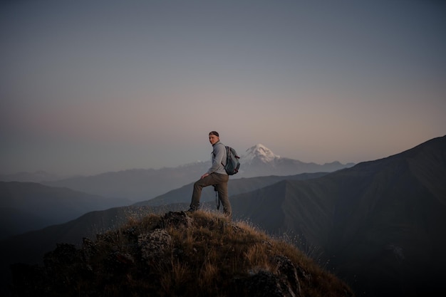 Joven excursionista caucásico parado en la cima de una montaña cubierta de hierba y admirando la naturaleza nublada