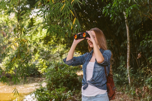 Joven excursionista mujer mirando a través de binoculares en el bosque