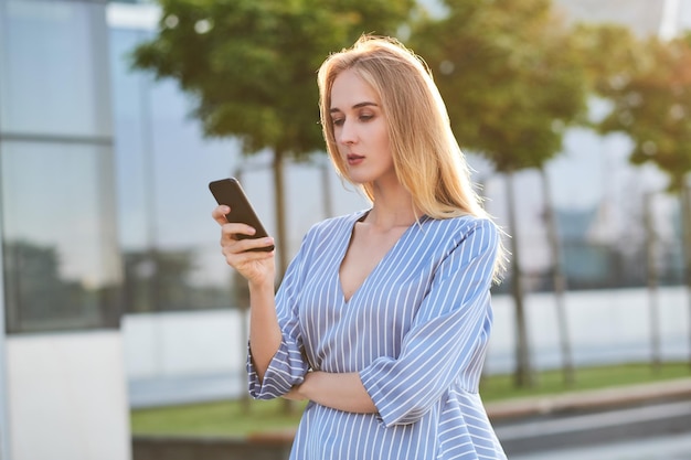 Una joven y exitosa mujer de negocios está esperando a su nueva pareja en la calle con un teléfono móvil en las manos.