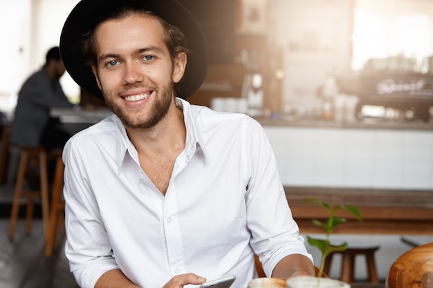 Joven exitoso con elegante sombrero relajándose solo en la acogedora cafetería durante el almuerzo, mirando con expresión feliz