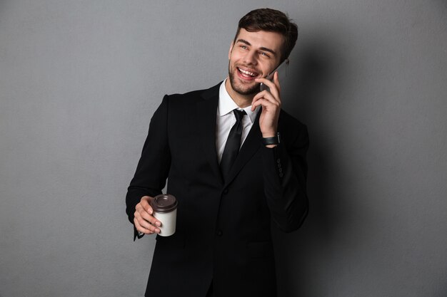Joven exitoso hombre de negocios hablando por teléfono móvil mientras sostiene una taza de café, mirando a un lado