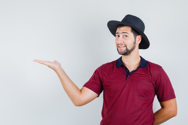 Joven extendiendo la palma a un lado en camiseta, sombrero y alegre. vista frontal.