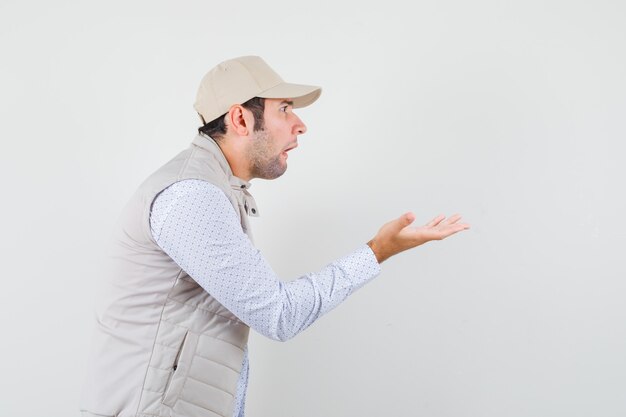 Joven extendiendo las palmas como sosteniendo algo en chaqueta y gorra beige y mirando sorprendido. vista frontal.