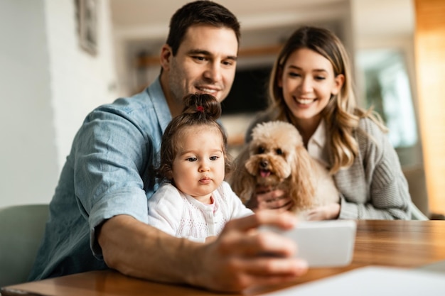 Joven familia feliz con un perro usando un teléfono inteligente mientras toma selfie en casa. El foco está en la chica.