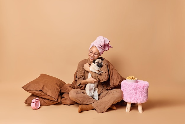 Foto gratuita una joven feliz abraza a su perro pug favorito expresa su amor y su pose de cuidado bajo una pose de manta en el dormitorio vestida con ropa de dormir aislada sobre un fondo marrón el dueño de la mascota se queda en una cama cómoda