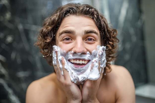 Joven y feliz afeitado con crema de afeitar en el baño.