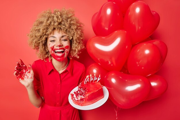 Una joven feliz y alegre untada con crema después de comer pastel contenta de celebrar el aniversario en un círculo de amigos viste ropa festiva aislada sobre un fondo rojo Concepto de ocasión festiva