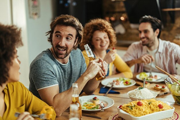 Joven feliz almorzando con amigos y bebiendo cerveza mientras se comunica con ellos en la mesa del comedor