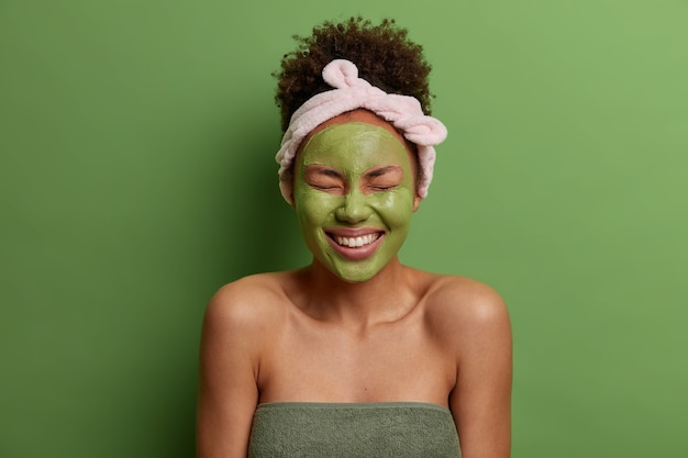 Joven feliz se aplica mascarilla de arcilla natural para reducir el acné, sonríe ampliamente, tiene dientes blancos perfectos, se ha peinado el cabello rizado, diadema, muestra los hombros desnudos, aislado en la pared verde