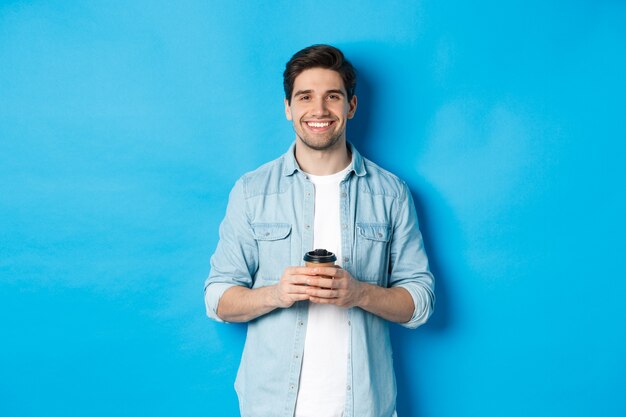 Joven feliz bebiendo café de la cafetería para llevar, sonriendo complacido, de pie contra el fondo azul.