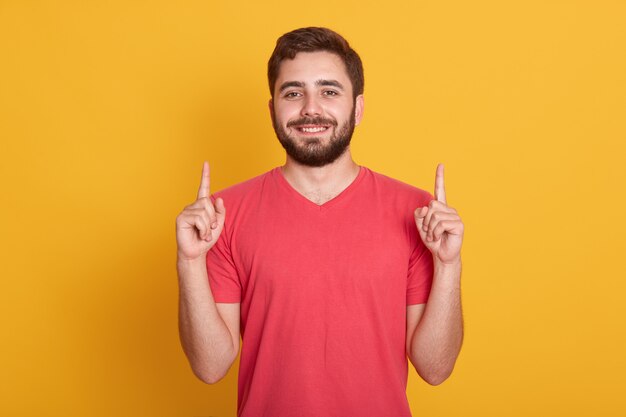 joven feliz con buen humor, posando aislado en amarillo, apuntando hacia arriba con los dedos índices, mirando sonriente. Copie espacio para publicidad o promoción.