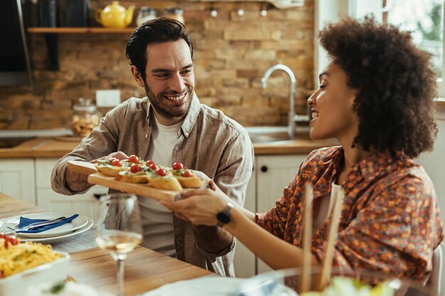 Joven feliz comunicándose con su novia afroamericana mientras le pasa la comida en la mesa del comedor