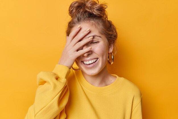 Una joven feliz cubre la mitad de la cara con sonrisas en las manos, sonríe alegremente, se ríe con dientes y expresa emociones positivas, cierra los ojos de la alegría aislada sobre el fondo amarillo, se encuentra en una pose relajada