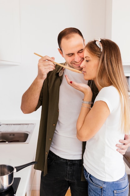 Foto gratuita joven feliz dejando que su esposa pruebe una sopa con una cuchara de madera en la cocina