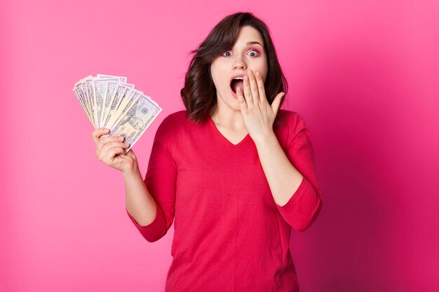 Joven feliz con dinero en mano, con la boca abierta, parece sorprendida. Chica morena gana en lotería. Suerte mujer vestida de jersey rojo quiere gastar su premio. Aislado en rosa