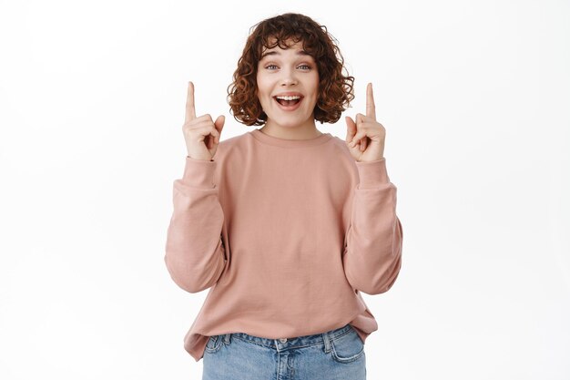 La joven feliz y emocionada se ve divertida, te sonríe mientras señala hacia arriba, muestra publicidad genial, descuentos promocionales hacia arriba en la pancarta, se para contra el fondo blanco.