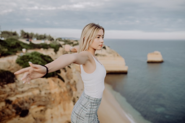 Joven feliz está sentada en lo alto y levanta los brazos con vista panorámica sobre la costa del océano