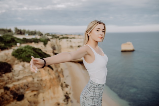 Joven feliz está sentada en lo alto y levanta los brazos con vista panorámica sobre la costa del océano