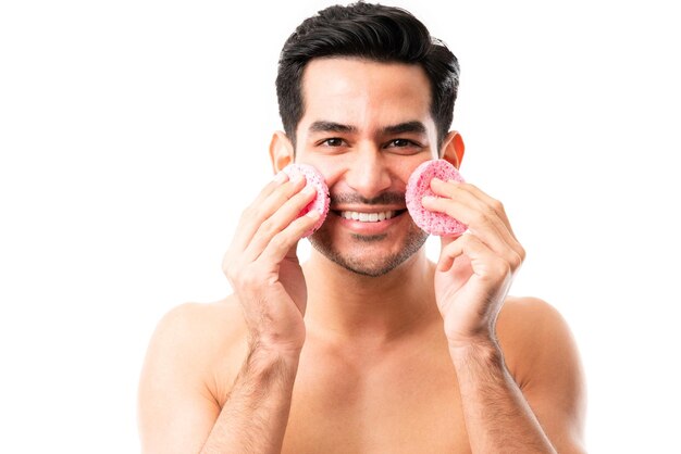 Un joven feliz limpia la piel con una esponja facial natural mientras hace contacto visual y sonríe con fondo blanco