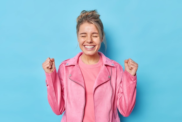 Una joven feliz y llena de alegría con el cabello peinado sonríe ampliamente mantiene los ojos cerrados anticipa los resultados levanta los puños apretados celebra los logros viste una chaqueta rosa aislada sobre un fondo azul