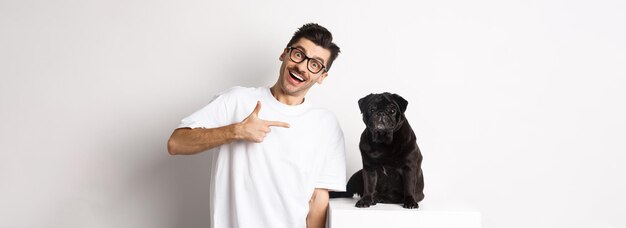 Un joven feliz mostrando a su lindo perro señalando con el dedo a un pug negro y sonriendo de pie sobre una ba blanca