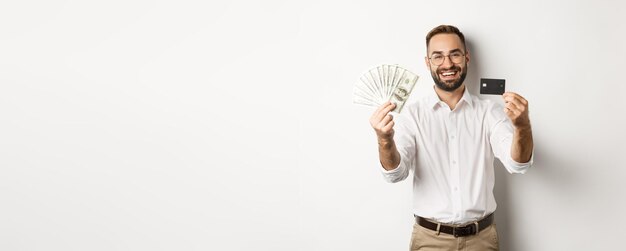 Joven feliz mostrando su tarjeta de crédito y dólares de dinero sonriendo satisfecho de pie sobre la espalda blanca