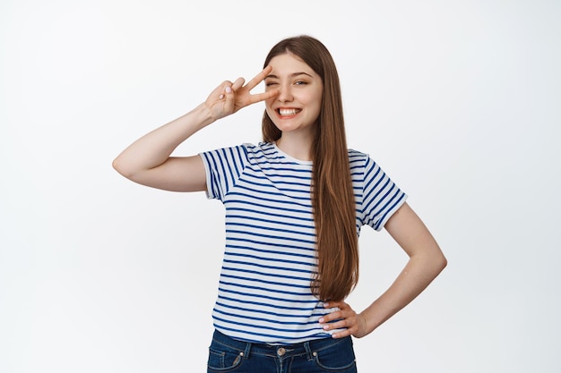 Una joven feliz muestra un gesto de disco en forma de v y sonríe a la cámara, de pie contra un fondo blanco con ropa informal.