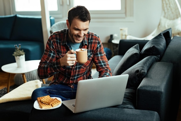 Joven feliz navegando por la red en una laptop mientras bebe café en casa