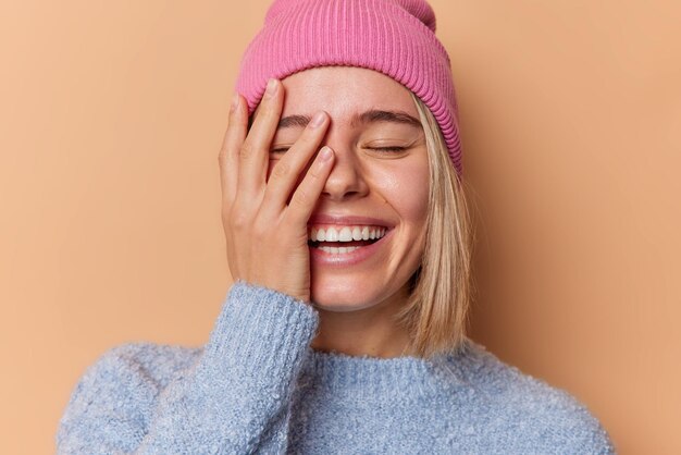 Una joven feliz y optimista hace que la cara de la palma sonría ampliamente, mantiene los ojos cerrados y está de buen humor, usa sombrero y suéter aislado sobre un fondo beige Concepto de emociones y sentimientos positivos