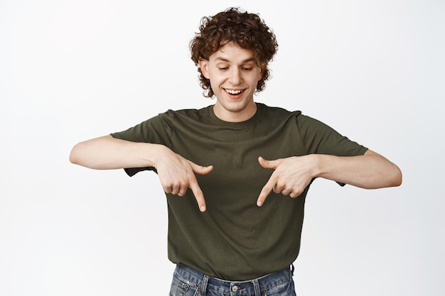 Un joven feliz con el pelo rizado apuntando y mirando hacia abajo sonriendo y riendo leyendo el anuncio del logotipo de pie sobre un fondo blanco